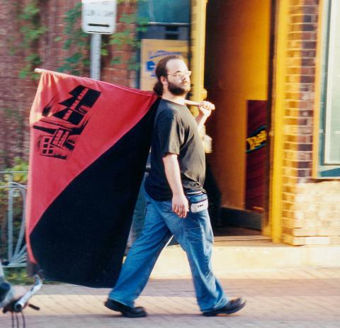 Photo : Nicolas porte un énorme drapeau rouge et noir sur lequel on disserne un dessin de domiciles. Jean, t-shirt noir, barbu, cheveux longs, lunettes.